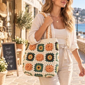 May include: A square crochet tote bag featuring a grid of alternating floral and geometric motifs in cream, dark green, and peach colors. The bag has two matching crochet shoulder straps and a structured, flat-bottomed rectangular shape.