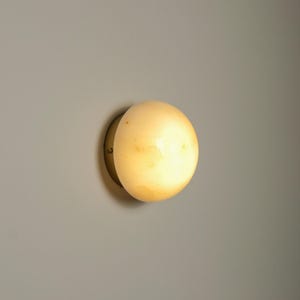 May include: A round wall-mounted light fixture featuring a spherical, translucent, cream-colored stone shade with natural veining patterns. The shade is secured to a circular, metallic brass-toned base that sits flush against the wall surface.