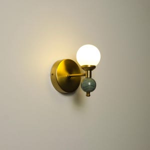 Könnte beinhalten: Eine Wandleuchte mit einer runden Rückplatte aus gebürstetem Messing, einem horizontalen Arm und einem vertikalen Stab. Die Leuchte verfügt über einen kugelförmigen weißen Glasschirm oben und eine kleinere, dekorative Kugel aus grauem Stein.