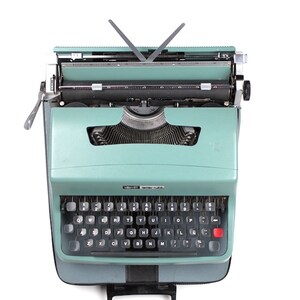Peut inclure: Une machine à écrire vintage verte avec un clavier noir et une touche de retour rouge. La machine à écrire est dans un étui de transport noir avec une poignée.