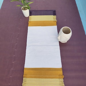Puede incluir: Un camino de mesa rectangular con un panel central blanco ancho flanqueado por rayas horizontales en amarillo mostaza, verde oliva claro y morado oscuro. Los extremos del camino de mesa están terminados con un detalle de flecos en morado oscuro.