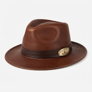 Puede incluir: Un sombrero fedora de cuero marrón con superficie texturizada, que presenta una banda marrón oscura alrededor de la copa. Una placa metálica dorada está fijada a la banda, mostrando el texto SPECIAL HAT con un icono de mano y detalles.