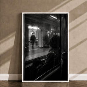 Può includere: Una stampa artistica da parete incorniciata in bianco e nero che raffigura una persona seduta all'interno di una carrozza ferroviaria che guarda fuori dal finestrino verso una banchina. La scena cattura una vista interna scura con una sagoma.