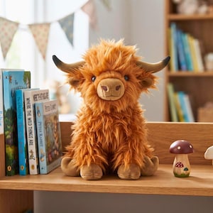 Puede incluir: Un juguete de peluche de vaca Highland con pelaje largo y desgreñado de color jengibre, cuernos oscuros curvados y un hocico suave de color tostado. El animal de peluche está sentado, con ojos oscuros y un pelaje texturizado de fibras sintéticas.