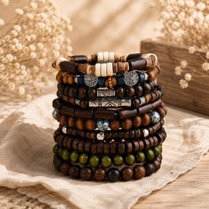 Könnte beinhalten: Ein Stapel aus neun Perlenarmbändern aus verschiedenen Materialien, darunter dunkelbraune Holzperlen, glatte olivgrüne Perlen, hellbeige zylindrische Perlen und silberfarbene Metallzwischenstücke mit Lebensbaum- und geometrischen Mustern.