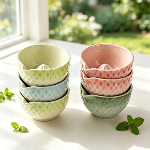 May include: A set of six ceramic citrus juicer bowls with a textured exterior and a central reamer. The bowls are arranged in two stacks of three, featuring pastel colours including light green, pale blue, soft pink, and a mottled sage green finish.
