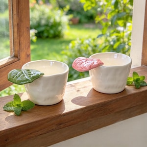May include: Two off-white ceramic cups with irregular, textured surfaces. One cup features a glossy green leaf-shaped handle, while the other has a glossy pink leaf-shaped handle. Both cups are positioned on a wooden surface with fresh mint leaves.