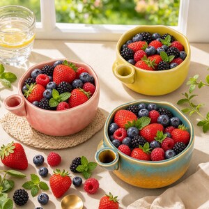 May include: Three ceramic bowls in yellow, pink, and a blue-to-yellow gradient, each featuring a small side handle. The bowls are filled with fresh strawberries, blackberries, raspberries, and blueberries, garnished with green mint leaves.