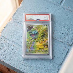 Könnte beinhalten: Eine bewertete 2023 Pokemon SV2a JP Bulbasaur Art Rare Sammelkarte in einer durchsichtigen Kunststoffhülle. Die Karte zeigt eine grüne Waldillustration mit dem Charakter Bulbasaur, 70 KP und dem sichtbaren Text Leech Seed.