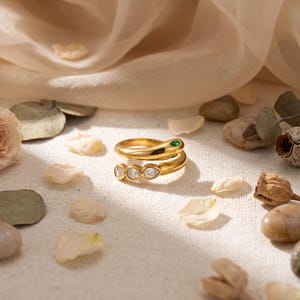 May include: A gold-toned bypass ring featuring a snake-inspired design with a small green stone for an eye and three clear, pear-shaped stones set along the lower band, creating a continuous, polished metallic wrap-around appearance.