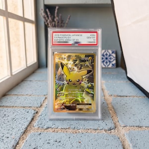 Op de afbeelding: Een Japanse Pokemon Pikachu EX ruilkaart uit 2016, 20e jubileumeditie, beoordeeld als GEM MT 10. De kaart toont een geel Pikachu-personage met bliksemeffecten, ingesloten in een doorzichtige, rechthoekige beschermende plastic houder.