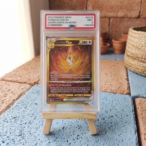 May include: A graded 2023 Pokemon SWSH Arceus VSTAR trading card from the Crown Zenith set, featuring a gold-toned illustration of the character. The card is encased in a clear plastic slab with a label at the top indicating a mint grade of 9.