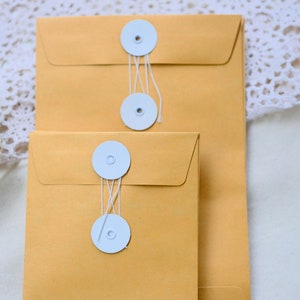 May include: Two brown envelopes with white string closures and white round buttons. The envelopes are stacked on top of each other.