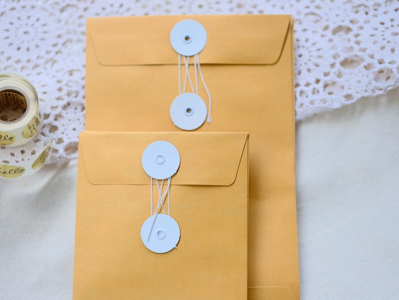 May include: Two brown envelopes with white string closures and white round buttons. The envelopes are stacked on top of each other.