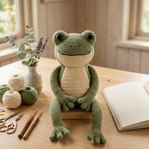 May include: A crocheted frog plush toy with olive green skin and a cream-colored belly, featuring large black eyes. Beside it are balls of yarn, two metal scissors, wooden crochet hooks, colorful stitch markers, and a small vase with greenery.