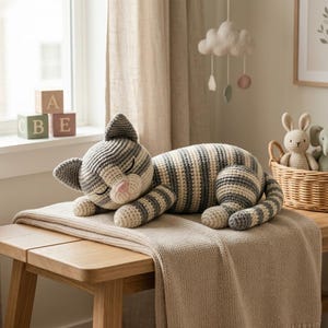 May include: A crocheted stuffed cat toy featuring horizontal stripes in shades of gray, white, and dark charcoal. The cat has a pink stitched nose, closed eyes, and pointed ears, depicted in a sleeping position with its head resting on its paws.