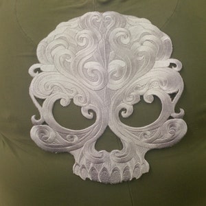 May include: A white embroidered skull design with intricate swirls and patterns on a green fabric background.