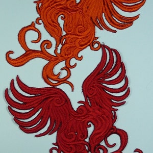 May include: Two embroidered phoenixes, one red and one orange, with intricate details and swirling patterns. The phoenixes are facing each other with their wings spread.
