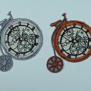 May include: Two embroidered iron-on patches depicting a bicycle with a clock face. One patch is silver with a black clock face and the other is orange with a black clock face.