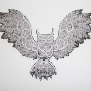 May include: A silver embroidered owl with intricate wing designs. The owl is facing forward with its wings spread out.