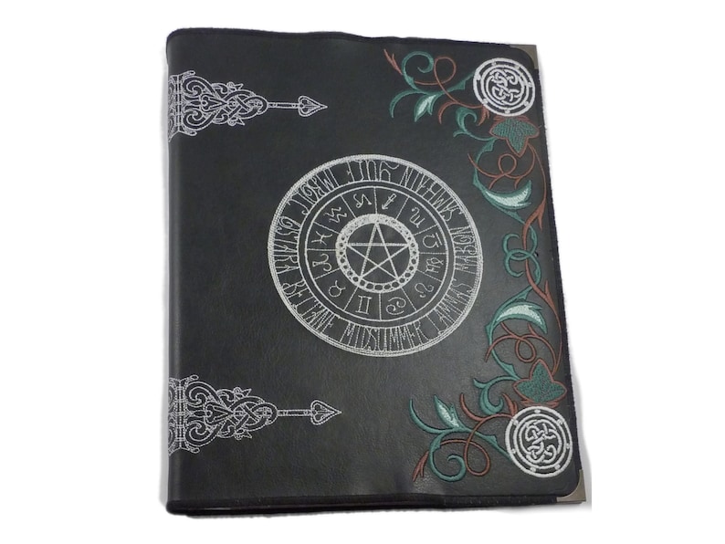 May include: Black leather journal featuring a silver pentagram and zodiac symbols, surrounded by green and red floral designs.