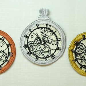 May include: Three embroidered iron-on patches in the shape of a pocket watch with intricate clockwork designs. The patches are in silver, gold, and copper.