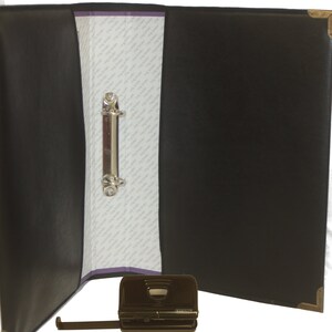 May include: Black leather binder with a silver ring mechanism and a black metal punch tool. The binder is open and the punch tool is lying on a white surface.