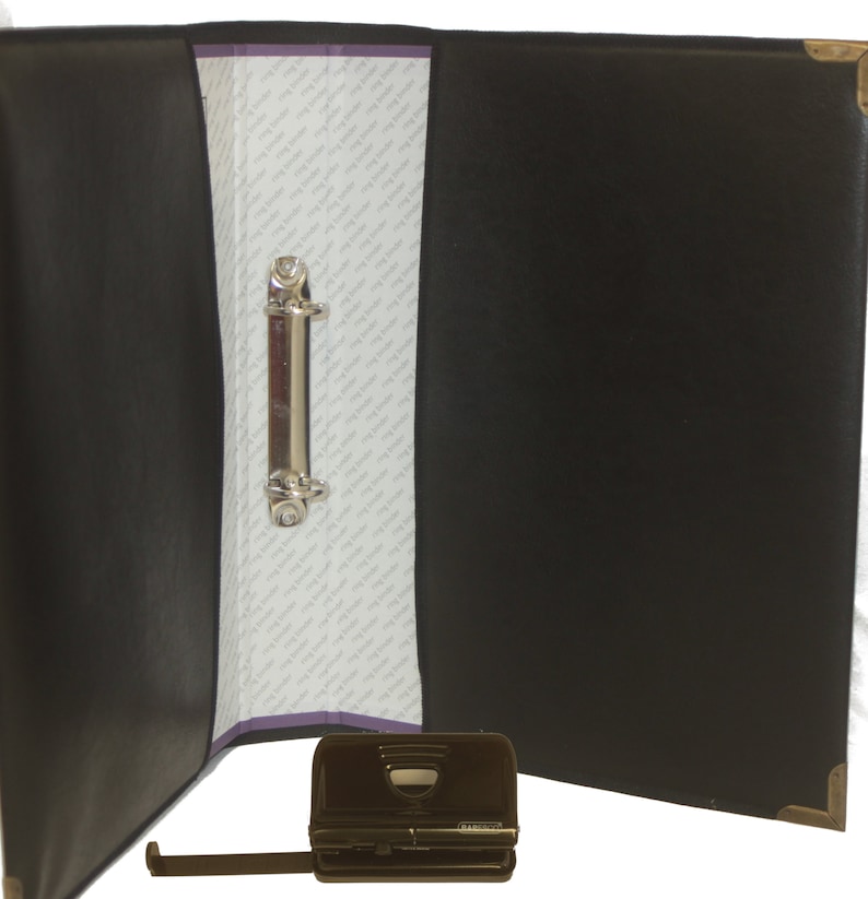 May include: Black leather binder with a silver ring mechanism and a black metal punch tool. The binder is open and the punch tool is lying on a white surface.