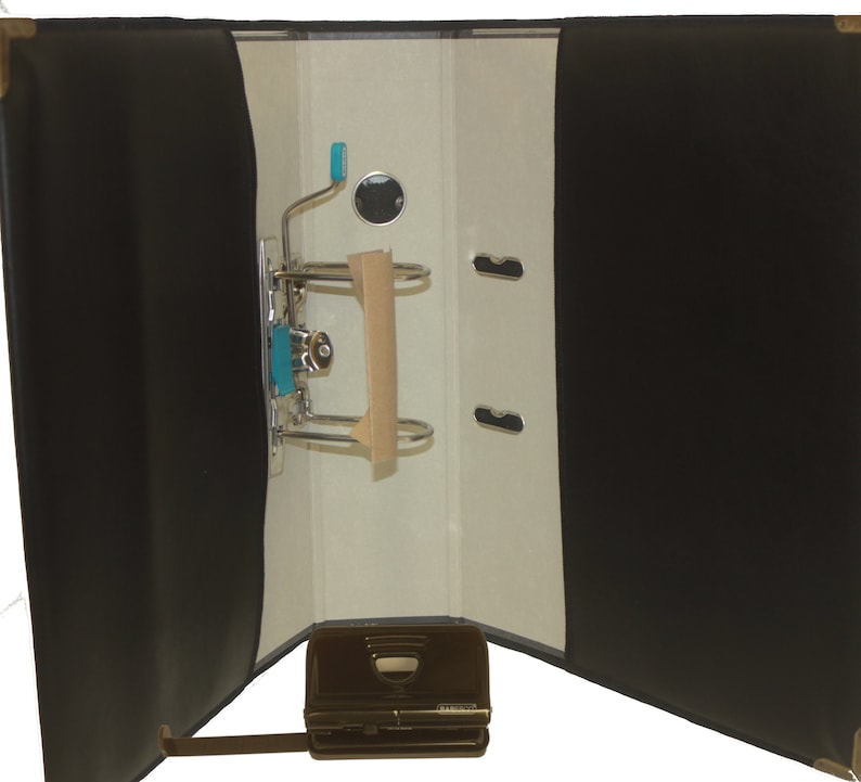 May include: Black leather binder with a metal ring mechanism and a built-in hole punch. The binder is open and the inside is white with a brown paper insert.