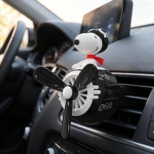 Puede incluir: Un ambientador de coche en blanco y negro con forma de pequeño avión con hélice. Una figura de perro blanco con gorro de aviador negro y bufanda roja está sentada encima. El cuerpo del avión presenta el texto SNOOPY y el número #01.