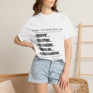 May include: A white short-sleeved t-shirt featuring black text that reads: THINGS I AM DOING RIGHT NOW, followed by a list: BEBOPPIN', SCHLEPPING, LOLLYGAGGING, CANOODLING, FLIBBERTIGIBBETING, and a partial line reading: (less to interrupt).