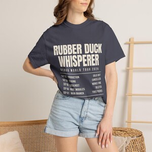 May include: A dark grey short-sleeve t-shirt featuring the white text RUBBER DUCK WHISPERER DEBUG WORLD TOUR 2026, followed by a list of dates and statuses including SEP 03 PRODUCTION SOLD OUT, SEP 04 STAGING CANCELLED, SEP 05 LOCALHOST WORKS FINE.