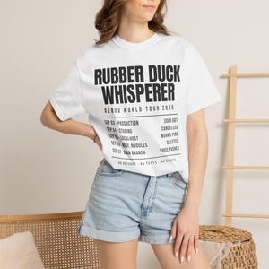 May include: A white short-sleeved t-shirt featuring black text that reads RUBBER DUCK WHISPERER DEBUG WORLD TOUR 2026, followed by a list of dates and statuses including SEP 03 PRODUCTION SOLD OUT, SEP 04 STAGING CANCELLED, and NO REFUNDS NO TESTS NO REGRETS.
