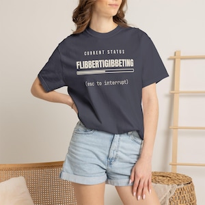 May include: A dark gray short-sleeved t-shirt featuring the white text CURRENT STATUS FLIBBERTIGIBBETING with a loading bar graphic and the text (esc to interrupt) printed on the front chest area of the garment.