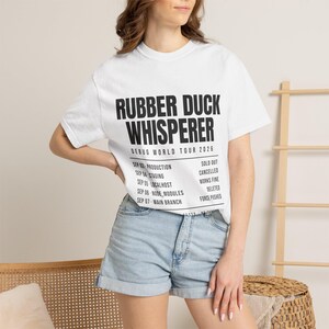 May include: A white short-sleeved t-shirt featuring black text that reads RUBBER DUCK WHISPERER, DEBUG WORLD TOUR 2026, and a list of dates and statuses including SEP 03 PRODUCTION SOLD OUT, SEP 04 STAGING CANCELLED, SEP 05 LOCALHOST WORKS FINE, SEP 06 NODE MODULES DELETED, SEP 07 MAIN BRANCH FORCE PUSHED.