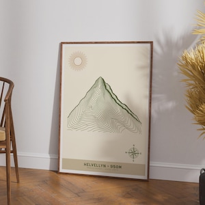 May include: A framed wall art print featuring a green topographic line map of Helvellyn mountain. The design includes a sun icon, a compass rose, and the text Helvellyn 950m on a cream background. The print is enclosed in a dark wood frame.