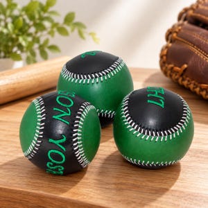 Puede incluir: Tres pelotas de béisbol con un diseño bicolor de paneles de cuero verde y negro. Cada pelota está cosida con hilo blanco. Una pelota muestra el texto bordado en verde "SON YOSH" y otra presenta el texto bordado en verde "LHT".