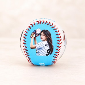 Könnte beinhalten: Ein weißer Baseball mit roten Nähten und einer hellblauen kreisförmigen Grafik auf der Vorderseite. Die Grafik zeigt eine Person mit langem dunklen Haar, die ein gestreiftes Trikot und einen Baseballhandschuh trägt, wobei die Nummer 9 zu sehen ist.