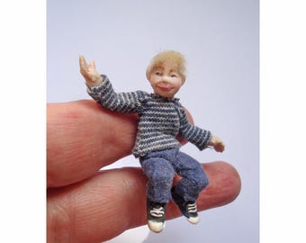 May include: A miniature doll with blonde hair wearing a grey and white striped long-sleeve shirt, blue pants, and black and white sneakers. The doll is posed with one arm raised. The text dollsbysallyafreeman is printed at the bottom of the image.