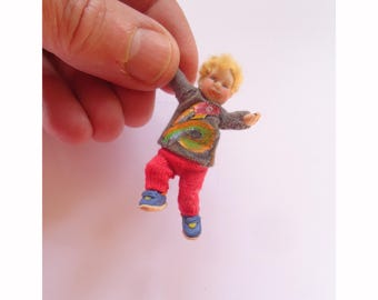 May include: A small, handmade doll of a child wearing a grey sweater with a colorful design and red pants. The doll is wearing blue shoes and has blonde hair.