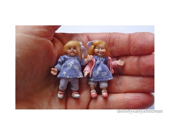 May include: Two small porcelain dolls wearing blue and pink dresses with white snowflake patterns. The dolls have blonde hair and are holding hands.