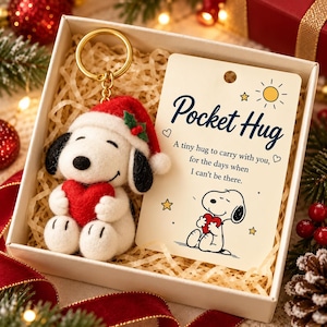 Puede incluir: Un llavero de perro de fieltro blanco con gorro de Papá Noel rojo y un corazón rojo, en una caja de regalo con una tarjeta que dice Pocket Hug, A tiny hug to carry with you, for the days when I can't be there, con una ilustración de Snoopy.