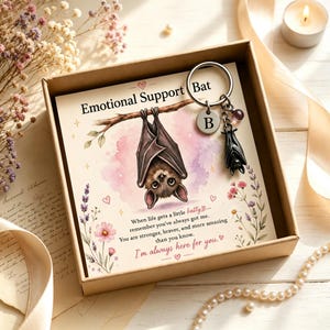 May include: A metal keychain featuring a small black bat charm, a round silver disc with the letter B, and a small purple bead, presented in a cardboard gift box with a card showing a bat illustration and the text Emotional Support Bat.