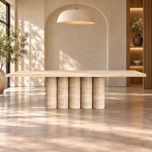 Puede incluir: Una mesa de comedor rectangular hecha de piedra travertino beige. La mesa presenta una superficie superior plana y lisa, apoyada por una base compuesta por cinco columnas cilíndricas verticales dispuestas en fila, todas elaboradas con la misma piedra texturizada.