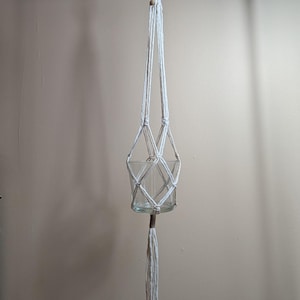 Peut inclure: Une suspension pour plante en macramé blanc avec un motif de cordes nouées retenant un récipient en verre transparent. La suspension est fixée à un anneau en bois et se termine par une longue frange décorative avec des perles en bois.