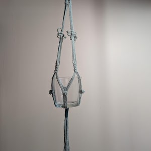 Peut inclure: Une suspension pour plante en macramé bleu clair avec des motifs de cordes nouées complexes et un récipient central en verre transparent. Le design comprend une boucle de suspension, des nœuds décoratifs et un long pompon suspendu.