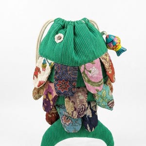 May include: A drawstring pouch shaped like a fish, featuring a green corduroy body with white pinstripes. The exterior is layered with various colorful floral and patterned fabric scales. A small wooden fish charm hangs from the drawstring.