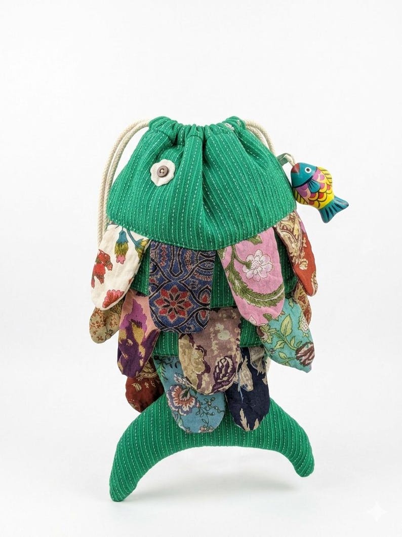 May include: A drawstring pouch shaped like a fish, featuring a green corduroy body with white pinstripes. The exterior is layered with various colorful floral and patterned fabric scales. A small wooden fish charm hangs from the drawstring.