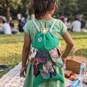 May include: A green drawstring backpack shaped like a fish, featuring a textured fabric body with a white flower accent and multiple overlapping fabric scales in various floral and geometric patterns in shades of red, blue, and brown.