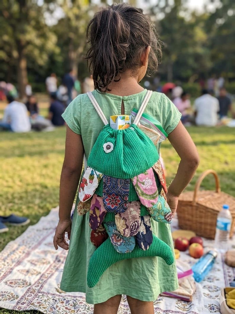 May include: A green drawstring backpack shaped like a fish, featuring a textured fabric body with a white flower accent and multiple overlapping fabric scales in various floral and geometric patterns in shades of red, blue, and brown.